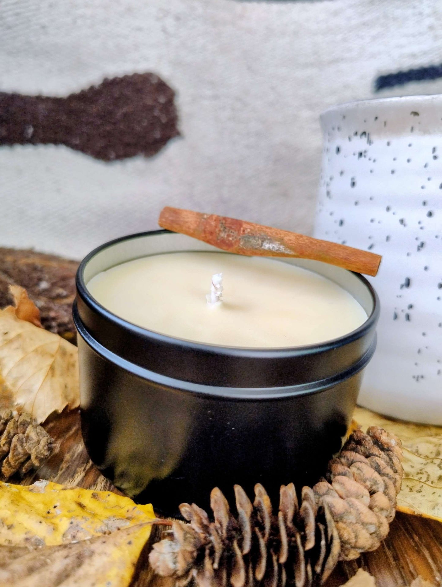Woodland Spice Candle in black tin with cinnamon stick lid surrounded by pine cones and autumn leaves