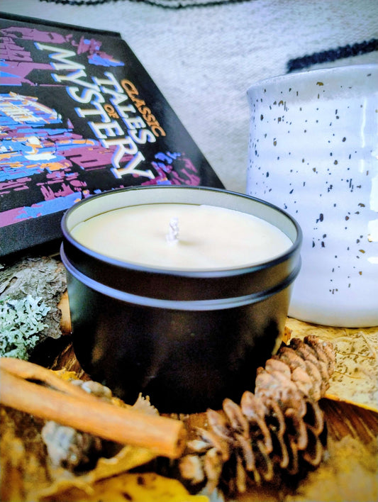 Woodland Spice Candle in black tin with pinecones, cinnamon stick, book, and speckled white ceramic cup on rustic surface