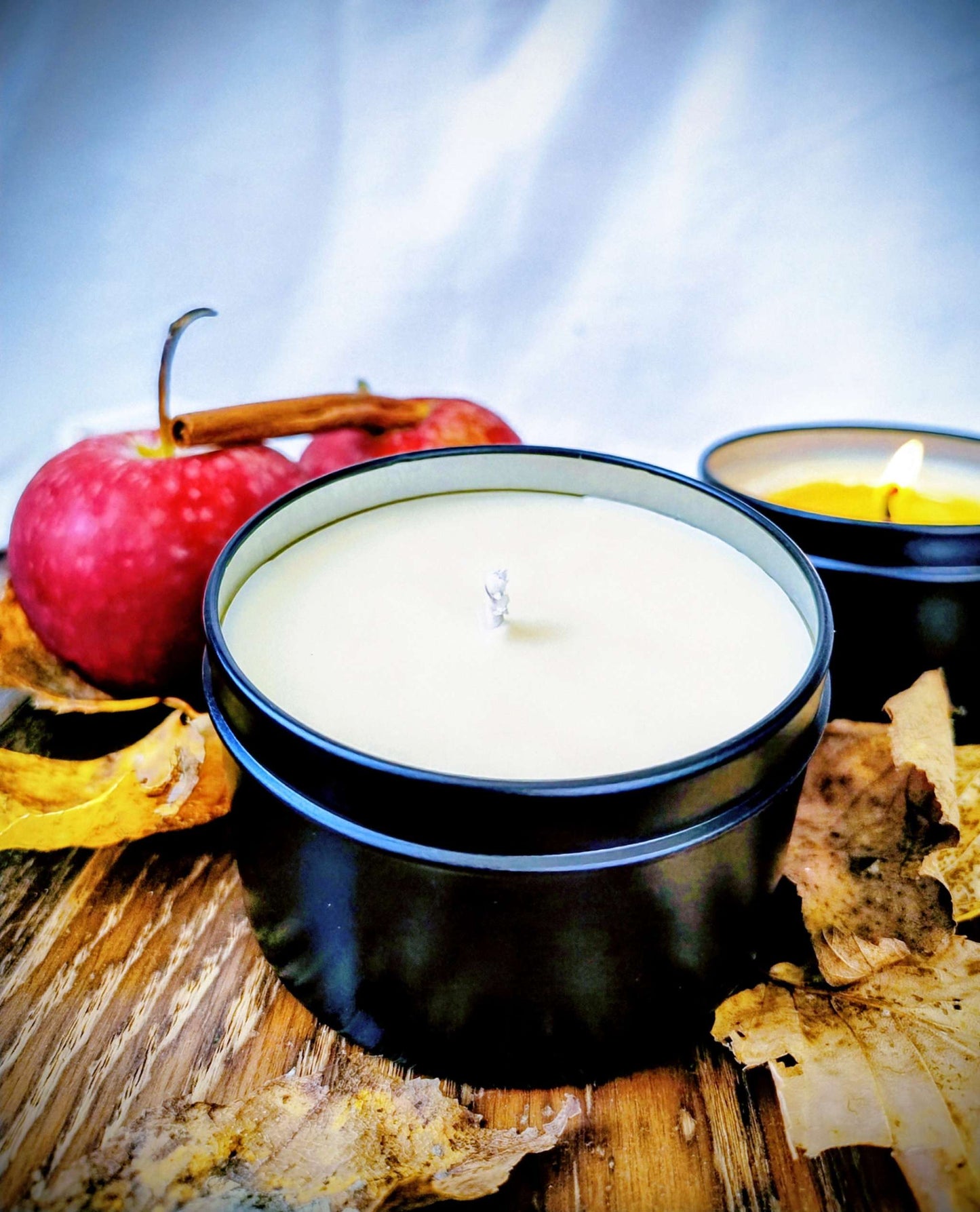Harvested Orchard Candle in black tin with apple, cinnamon stick, and dried leaves on wooden surface, evoking autumn warmth and citrus-spice fragrance