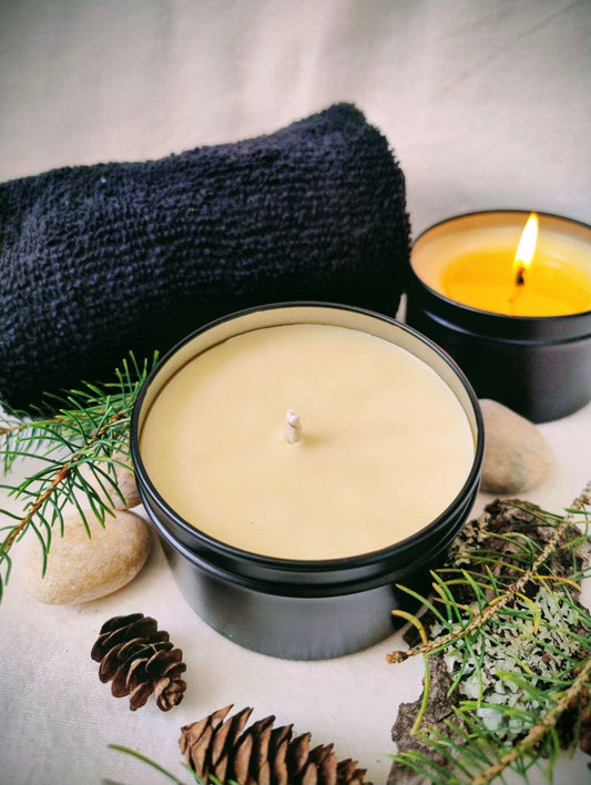 Forest Rain Candle in black tin with natural soy beeswax, surrounded by pinecones, stones, evergreen sprigs, and a folded dark towel, one candle lit in the background