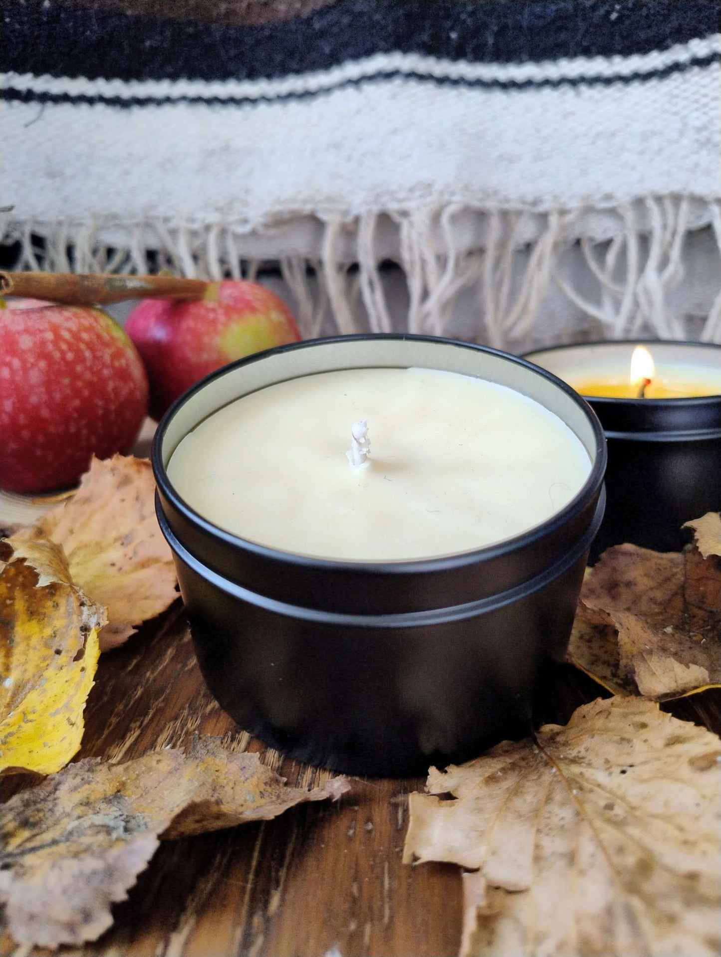 Harvested Orchard Candle in black tin surrounded by autumn leaves and apples on wooden surface