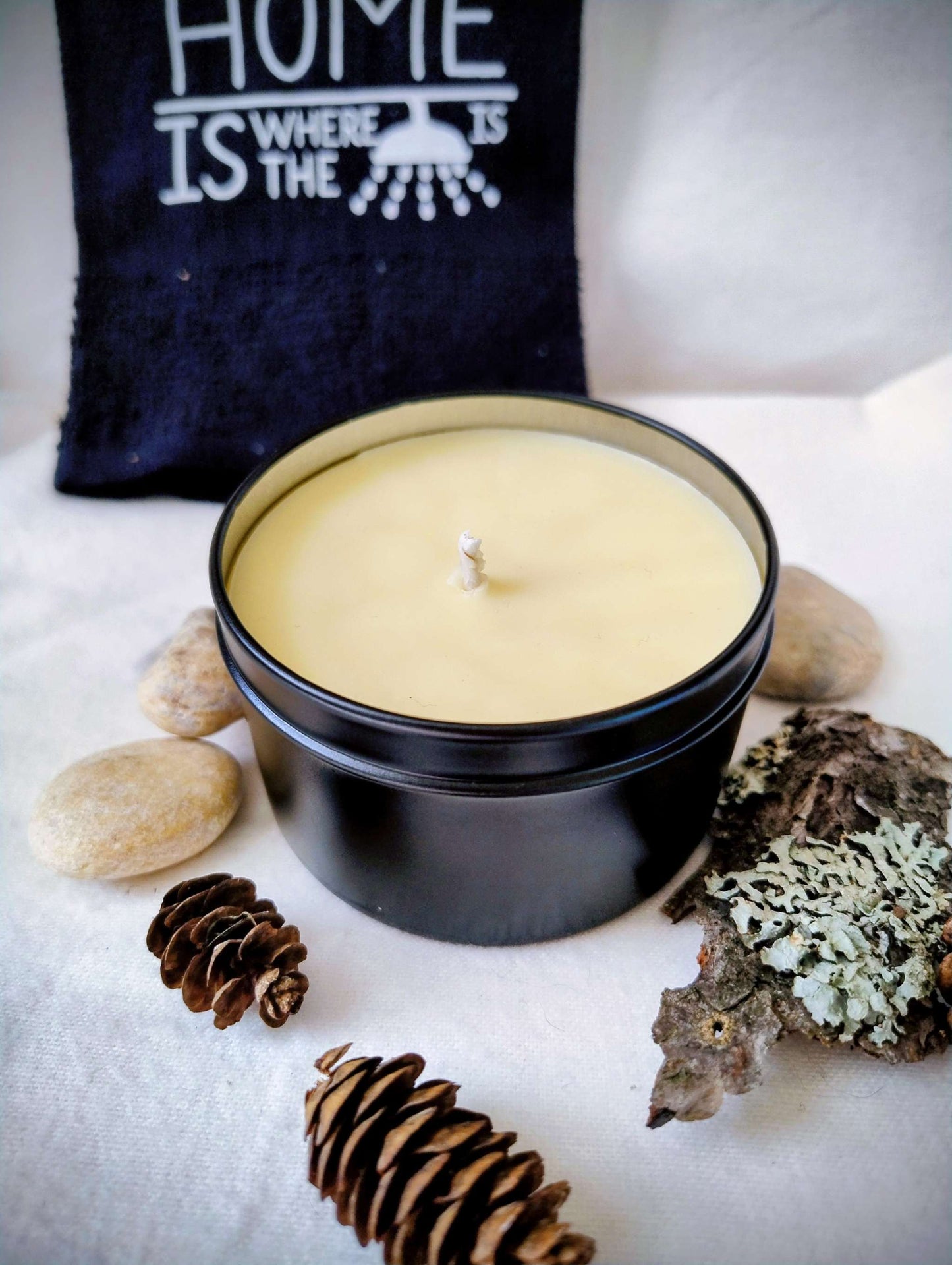 Forest Rain Candle in black tin with pine cones, lichen-covered bark, and smooth stones, evoking misty woodland freshness and earthy calm.