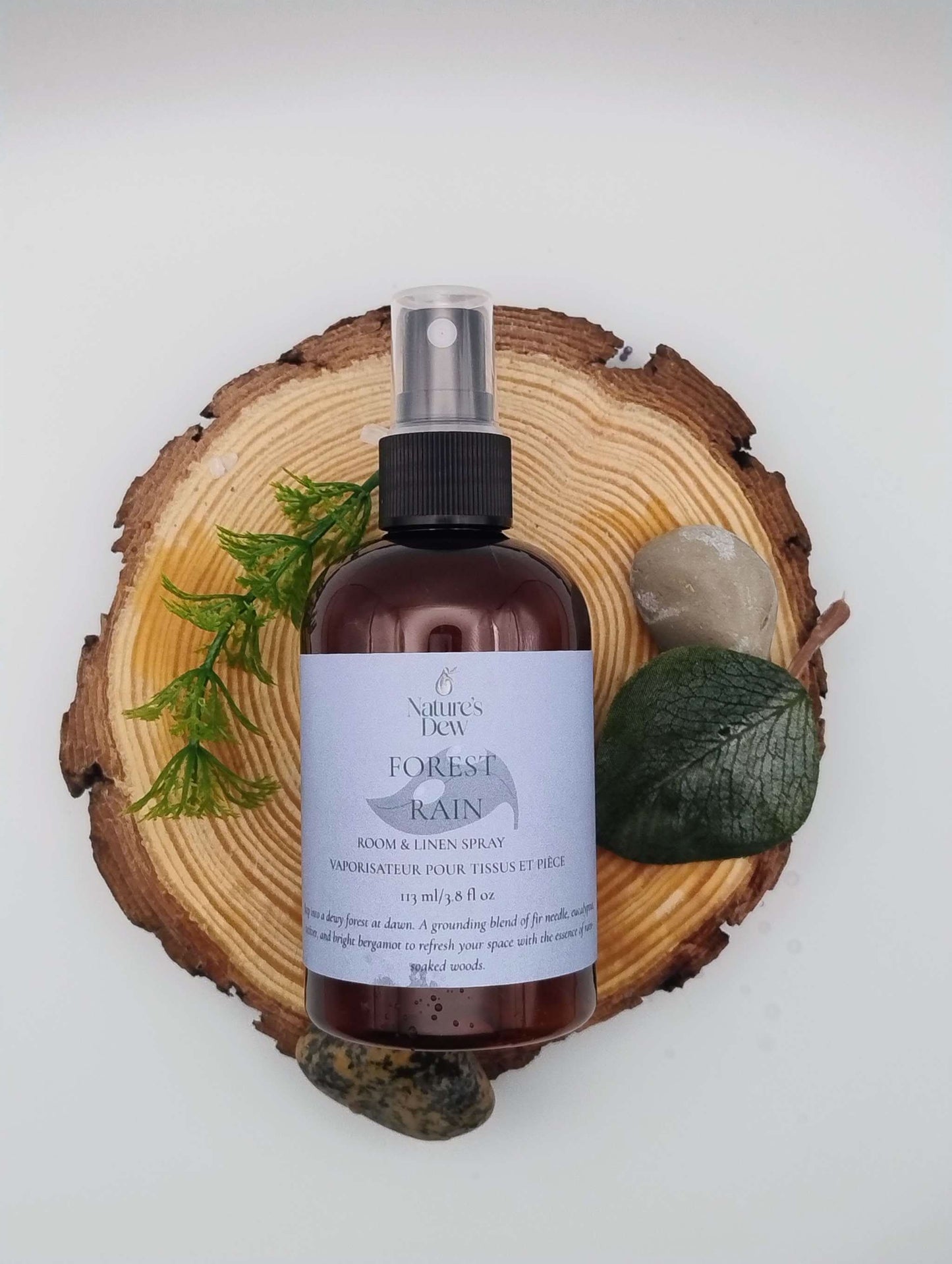 Forest Rain Room & Linen Spray bottle displayed on a wooden slice with green leaves and stones.